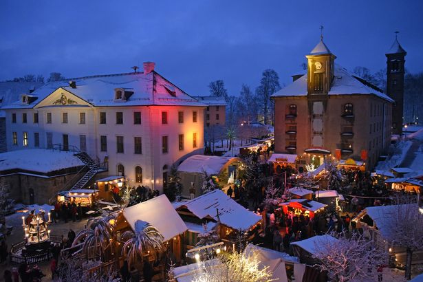 Auf dem historisch-romantischen Weihnachtsmarkt auf der Festung Königstein stehen verschiedene Schaubuden, deren Dächer mit leichtem Schnee bedeckt sind. Links ist eine Pyramide zu sehen. Der Platz mit dem Weihnachtsmarkt ist von mehreren Gebäuden umschlossen. Vor den Schaubuden stehen Menschen und schauen sich die Waren an, die verkauft und angeboten werden.