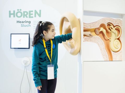 Eine Frau steht im Ausstellungsraum "Hören" des Deutschen Hygienemuseums Dresden. Sie greift mit ihrer linken Hand in ein großes Ohr. Dahinter sieht man die einzelnen Teile.