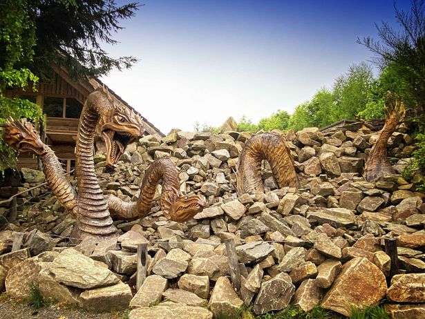 Ein überlebensgroßer Drache aus Holz windet sich durch aufgestapelte Steine. Im Hintergrund ist eine Holzhütte angedeutet. Es ist Holzkunst im Naturraum.