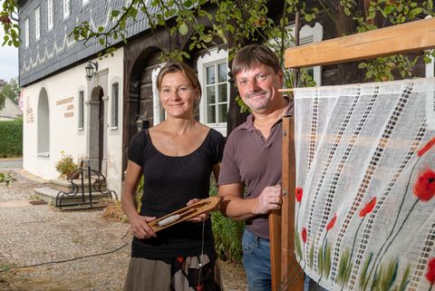 Ein Mann und eine Frau stehen an einem gewebten Stoff mit Blumenmuster. Sie sind am Haus Spinnwebe in Obercunnersdorf. 