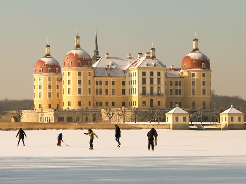 Das Jagdschloss Moritzburg im Winter ist abgebildet. Vor dem Schloss laufen Leute Schlittschuh auf dem zugefrorenen See.