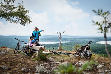 Eine Familie mit zwei Kindern macht Pause auf einer Bank auf der Blockline. Sie haben eine gute Aussicht auf das Erzgebirge mit Wäldern und Seen. Der Vater hält die Tochter auf dem Arm und zeigt ihr etwas.