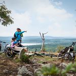 Eine Familie mit zwei Kindern macht Pause auf einer Bank auf der Blockline. Sie haben eine gute Aussicht auf das Erzgebirge mit Wäldern und Seen. Der Vater hält die Tochter auf dem Arm und zeigt ihr etwas.