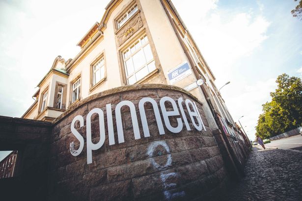 Ein Gebäude im Hintergrund. Davor steht eine Mauer mit dem Schriftzug "Spinnerei" versehen. Die Steinmauer ist mit Graffiti bemalt.