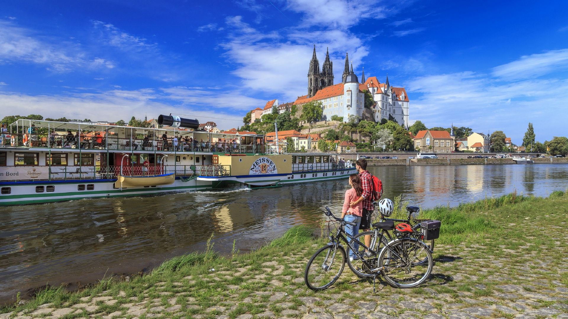 Ein Paar steht mit den Fahrrädern am Elberadweg in Meißen. Sie schauen auf die Elbe, wo eine Dampfschiff liegt und auf die Albrechtsburg von Meißen. 