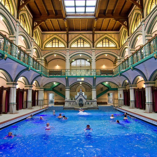 Besucher schwimmen in einem Becken des Johannisbad in Zwickau. Die Wände sind verziert und ein prunkvolles Geländer umrahmt das Ganze. 