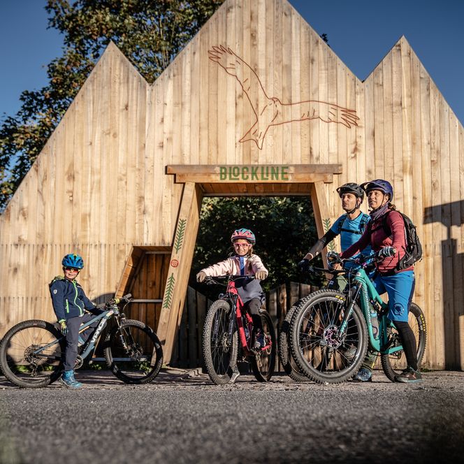 Eine Familie steht mit ihren Mountainbikes vor einem Eingangstor aus Holz. 