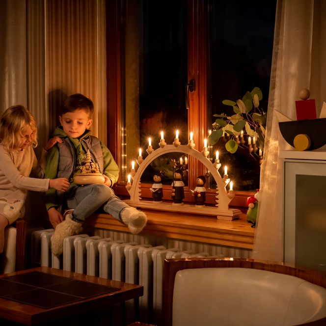 Zwei Kinder sitzen auf einer Heizung und schauen sich eine Spieluhr aus Holz an. Auf dem Fensterbrett steht ein Schwibbogen mit leuchtenden Kerzen. Draußen vor dem Fenster ist es dunkel.