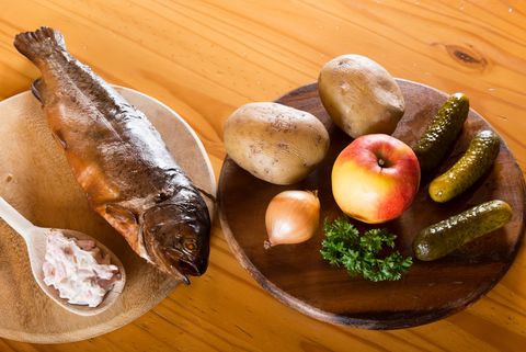 Eine geräucherte Forelle liegt auf einem Holzbrett. Daneben steht eine Platte mit Kartoffeln, Gurken und einem Apfel. 