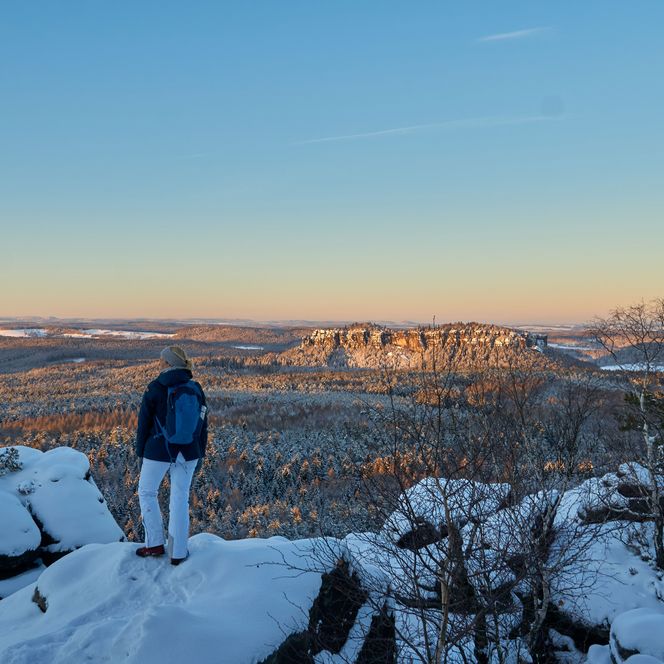 Eine Wanderin steht auf den schneebedeckten Felsen des Elbsandsteingebirges und blickt über die Winterlandschaft.
