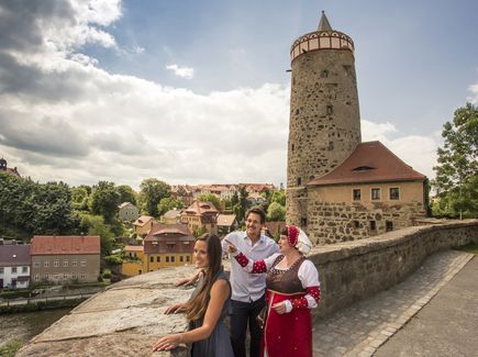 Es ist ein Stadtrundgang in Bautzen abgebildet. Ein Pärchen steht zusammen mit einer Frau in traditioneller Tracht an einer Mauer. Die Frau zeigt auf etwas, was in der Ferne liegt. Im Hintergrund ist ein Turm und hinter der Mauer befinden sich einzelne Gebäude, ein Fluss und Bäume. 