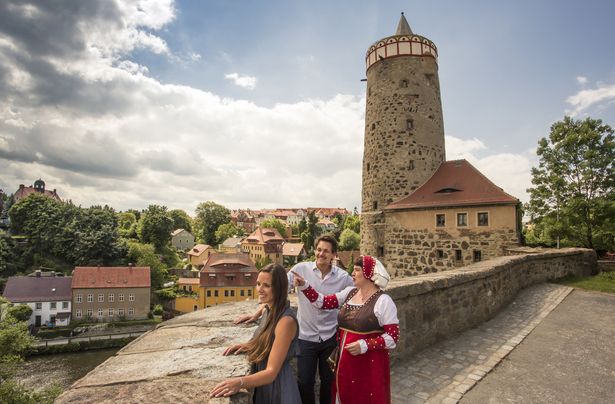 Es ist ein Stadtrundgang in Bautzen abgebildet. Ein Pärchen steht zusammen mit einer Frau in traditioneller Tracht an einer Mauer. Die Frau zeigt auf etwas, was in der Ferne liegt. Im Hintergrund ist ein Turm und hinter der Mauer befinden sich einzelne Gebäude, ein Fluss und Bäume. 