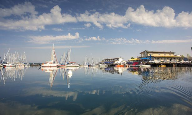 Eine Anlegestelle mit einem Haus und Steg auf der rechten Seite. Links sieht man Segelschiffe und Boote auf dem Wasser des Cospudener Sees.