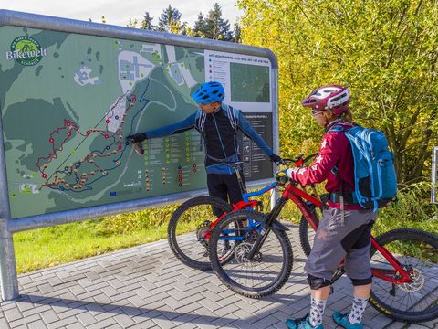 Ein Mann und eine Frau stehen mit ihren Mountainbikes an einer Tafel mit einer Karte. Sie sind in der Bikewelt Schöneck.