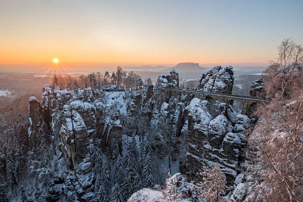 Das Bild zeigt die Aussicht auf die Tafelberge der Sächsischen Schweiz. Sie sind mit Schnee bedeckt. Die Sonne steht kurz oberhalb des Horizonts. Es herrscht heimelige Winterstimmung. Es sind keine Menschen zu sehen, nur Natur.