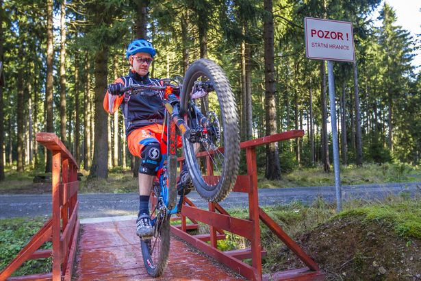 Mountainbiker fährt nur auf dem Hinterrad über eine Brücke im Wald. Er ist auf dem MTB-Track Stoneman Miriquidi unterwegs. Er trägt Helm und MTB-Ausrüstung.