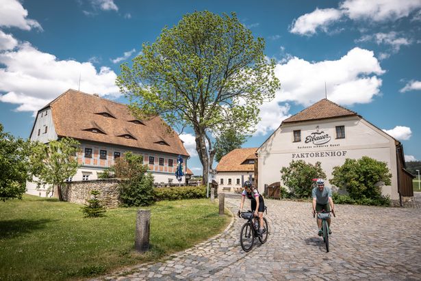 Zwei Gravelbike-Fahrer vor einem Drei-Seiten-Hof. Beide fahren auf gepflastertem Untergrund. Die Sonne scheint. Im Hintergrund ist ein großer Laubbaum zu sehen.
