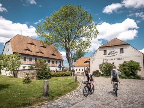 Zwei Gravelbike-Fahrer vor einem Drei-Seiten-Hof. Beide fahren auf gepflastertem Untergrund. Die Sonne scheint. Im Hintergrund ist ein großer Laubbaum zu sehen.