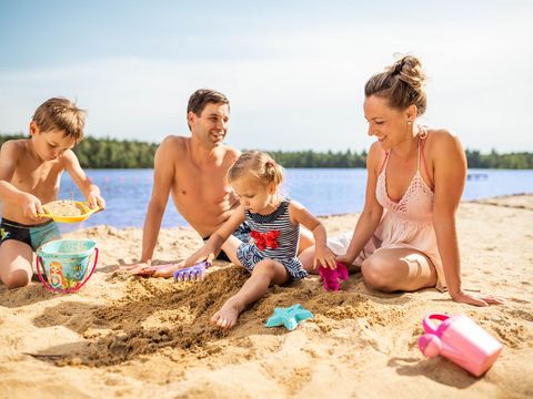 Eine Familie sitzt am Strand, die Kinder graben im Sand. Dahinter ist ein See.