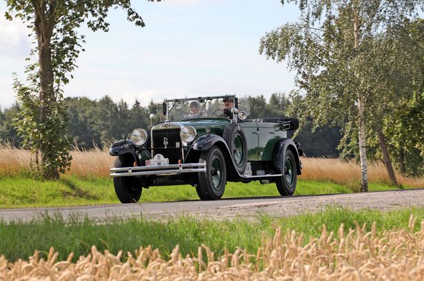 Oldtimer bei einer Ausfahrt aus dem August Horch Museum. Ein Oldtimer ist auf einer kleinen Straßen unterwegs. In dem Oldtimer sitzt ein älteres Pärchen. Um die Straße herum sind Wiesen, einzelne Bäume und im Hintergrund ein Wald.