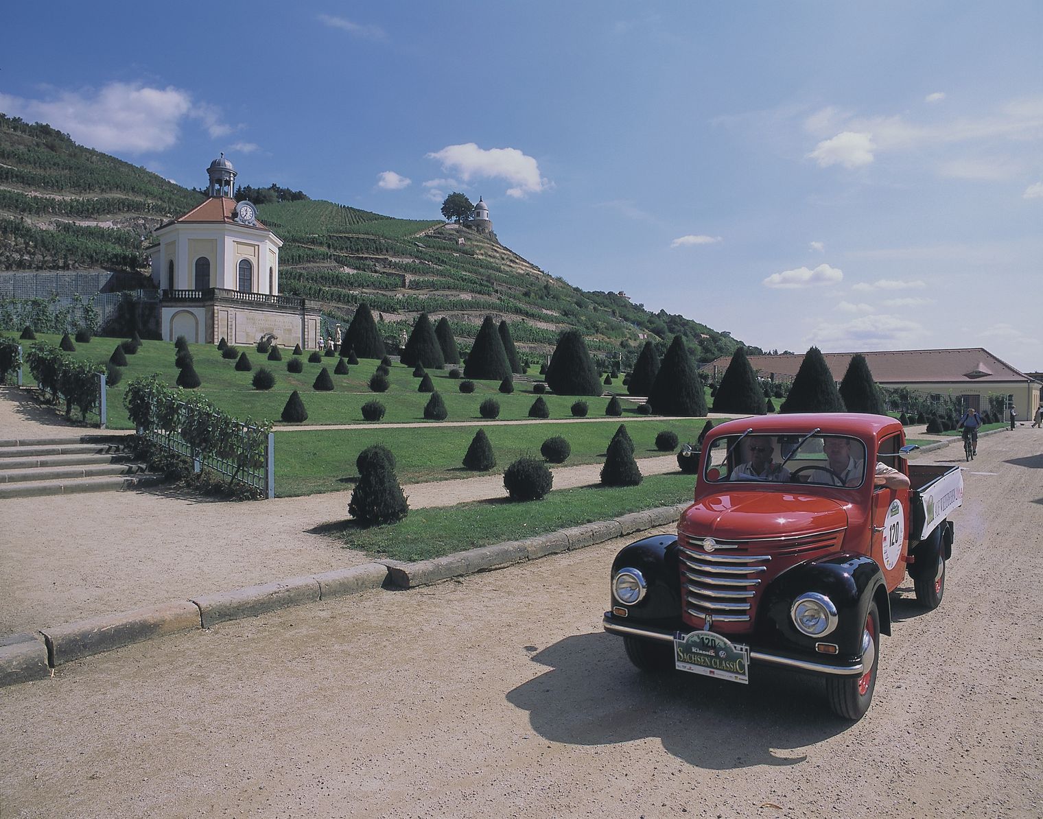 Ein Oldtimer steht vor dem Schloss Wackerbarth. Zwei Leute sitzen im Auto und im Hintergrund befinden sich Weinberge.