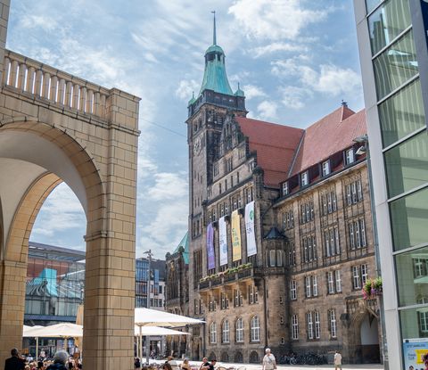 Marktplatz einer größeren Innenstadt gesäumt von historischen und modernen Gebäuden. Ein Mensch läuft uns den gepflasterten Bodenbelag.