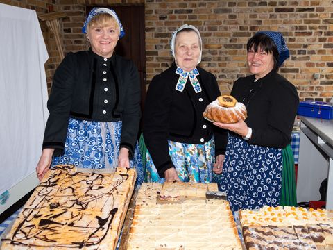 Drei Frau in sorbischen Trachten stehen vor großen Blechen mit Kuchen. Sie sind vom Hof-Verein Schleife. 