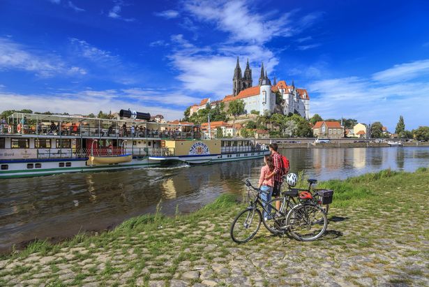 Ein Paar steht mit den Fahrrädern am Elberadweg in Meißen. Sie schauen auf die Elbe, wo eine Dampfschiff liegt und auf die Albrechtsburg von Meißen. 