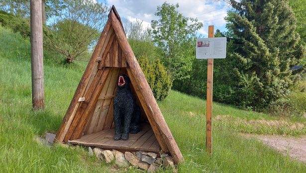 Im Bild ist eine dreieckig, spitz zulaufende Hütte zu sehen, deren Front offen ist. Darin sitzt ein schwarzes Fabelwesen, welches sein Mail aufreißt. Im Hintergrund des Bildes ist Natur zu sehen. Die Wiesen grünen. Es ist Sommer.