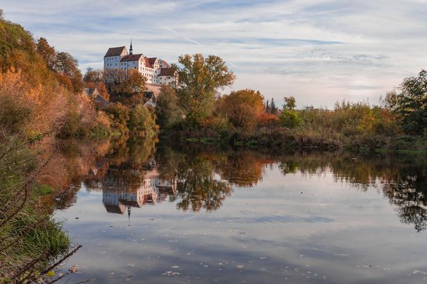 Colditz Schloss Ein weißes Schloss mit roten Dächern steht auf einer Anhöhe am Fluss. Die Bäume um das Schlos verfärben sich schon herbstlich und das Schloss spiegelt sich im Wasser.