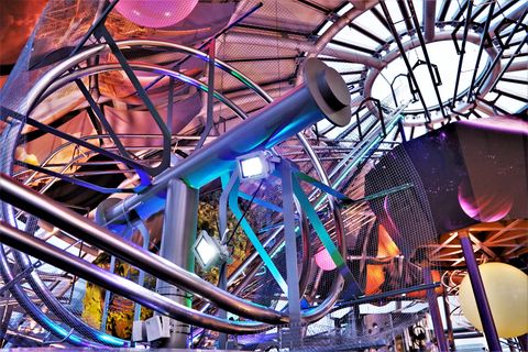 Eine farbenfrohe und futuristische Installationen mit metallischen Rohren und Lichtern in einem künstlerischen Raum, der an eine Weltraumumgebung erinnert. Im Hintergrund sind schimmernde, planetenähnliche Objekte und eine Lichtinstallation sichtbar, die eine dynamische und moderne Atmosphäre schaffen.