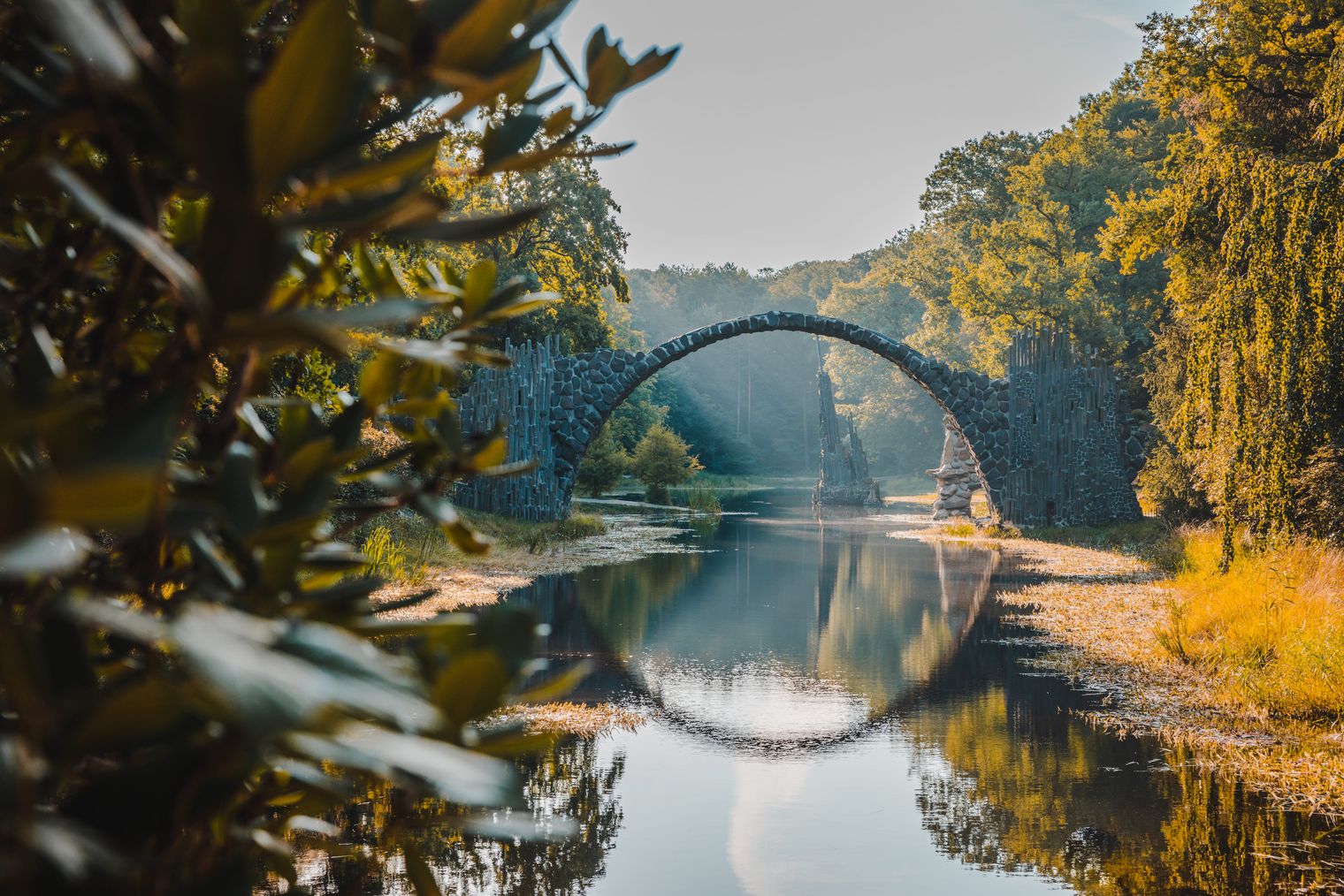 Halbrunde Rücke aus Stein im Hintergrund, die über Wasser reicht. Am Rand des Wasserfläche stehen Bäume und andere Pflanzen.