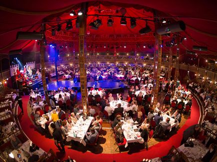 Das Bild zeigt in der Froschperspektive einen Varieté-Saal von Innen. An größeren Tischen sitzen Personen und nehmen das Abendessen ein. In der Mitte des Raums ist eine Bühne aufgebaut. Die Tische sind in einem Kreis angeordnet. Rings um den Kreis herum liegt roter Teppich aus.