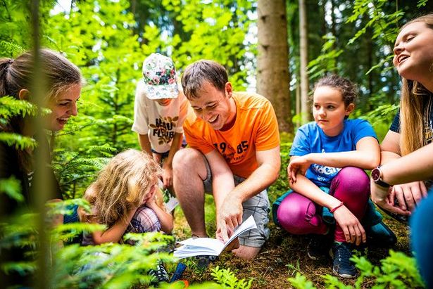 Kinder hocken in einem Wald bei einem KiEZ. Ein Mann zeigt ihnen etwas in einem Buch.