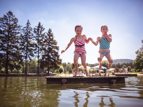Zwei Mädchen springen von einem Steg ins Wasser im Trixi Ferienpark. Dahinter sind Bäume.