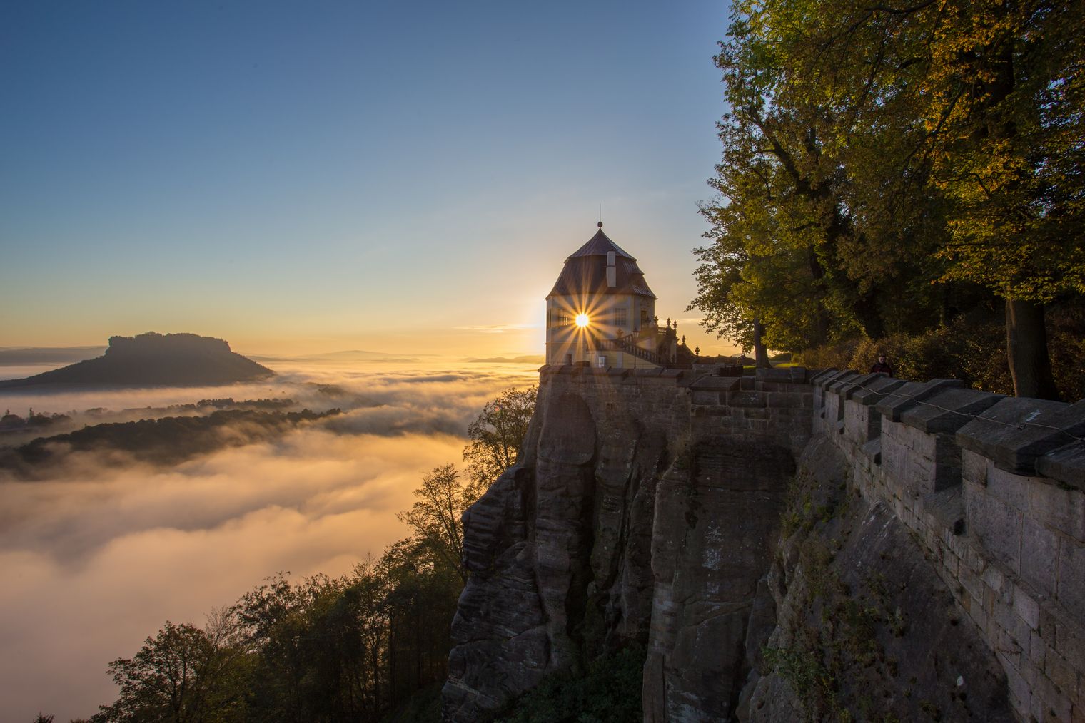 Eine Burg auf einem Berg. Unterhalb ist Nebel. Die Sonne geht gerade unter. 