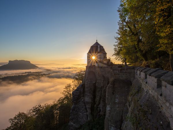 Eine Burg auf einem Berg. Unterhalb ist Nebel. Die Sonne geht gerade unter.