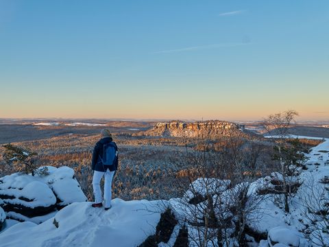 Eine Wanderin steht auf den schneebedeckten Felsen des Elbsandsteingebirges und blickt über die Winterlandschaft.