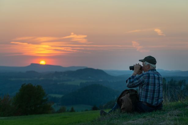 Das Bild wurde in Saupsdorf in der Sächsischen Schweiz aufgenommen. Es zeigt einen Mann, der auf einer Wiese sitzt und mit dem Fernglas in die Ferne schaut. Die Wiese ist auf einer Erhebung und in der Umgebung sind Hügel und Wälder zu sehen. Die Sonne geht gerade hinter einem Hügel unter.