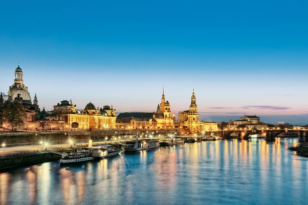 Am Ufer der Elbe befinden sich die prächtigen Gebäude der Dresdner Altstadt. Es ist Abend. Der Himmel färbt sich dunkel und überall leuchten Laternen.