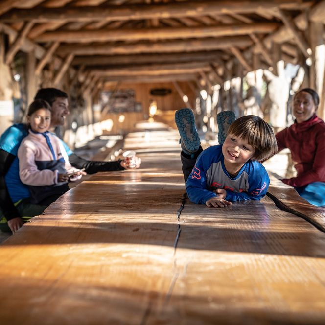 Eine Familie sitzt an einem langen Holztisch und der Sohn liegt auf dem Tisch und freut sich.
