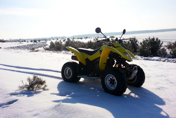 Das Bild zeigt ein gelbes Quad, welches auf Schnee-bedecktem Untergrund steht. Auf dem Quad sitzt keine Person. Im Schnee sind die Anfahrtsspuren der Reifen des Quads zu sehen. Das motorisierte Vierrad steht am Ufer eines Sees. Es herrscht Winterstimmung. 