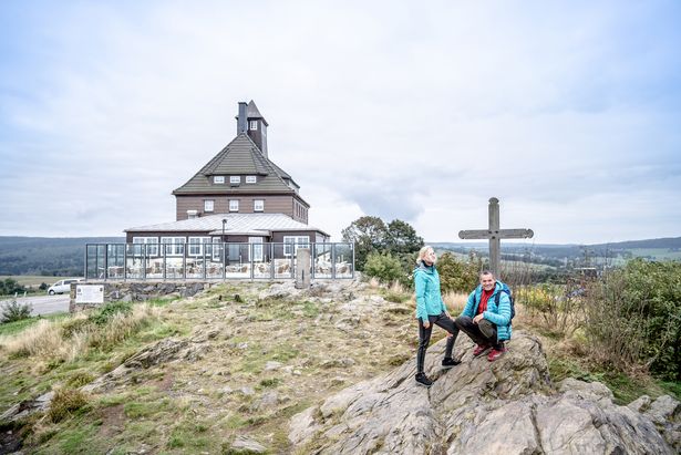 Auf dem Bild ist der Kammweg und der Schwartenberg abgebildet. Zwei Wanderer befinden sich am Gipfelkreuz. Hinter ihnen ist ein Gebäude mit Terrasse.