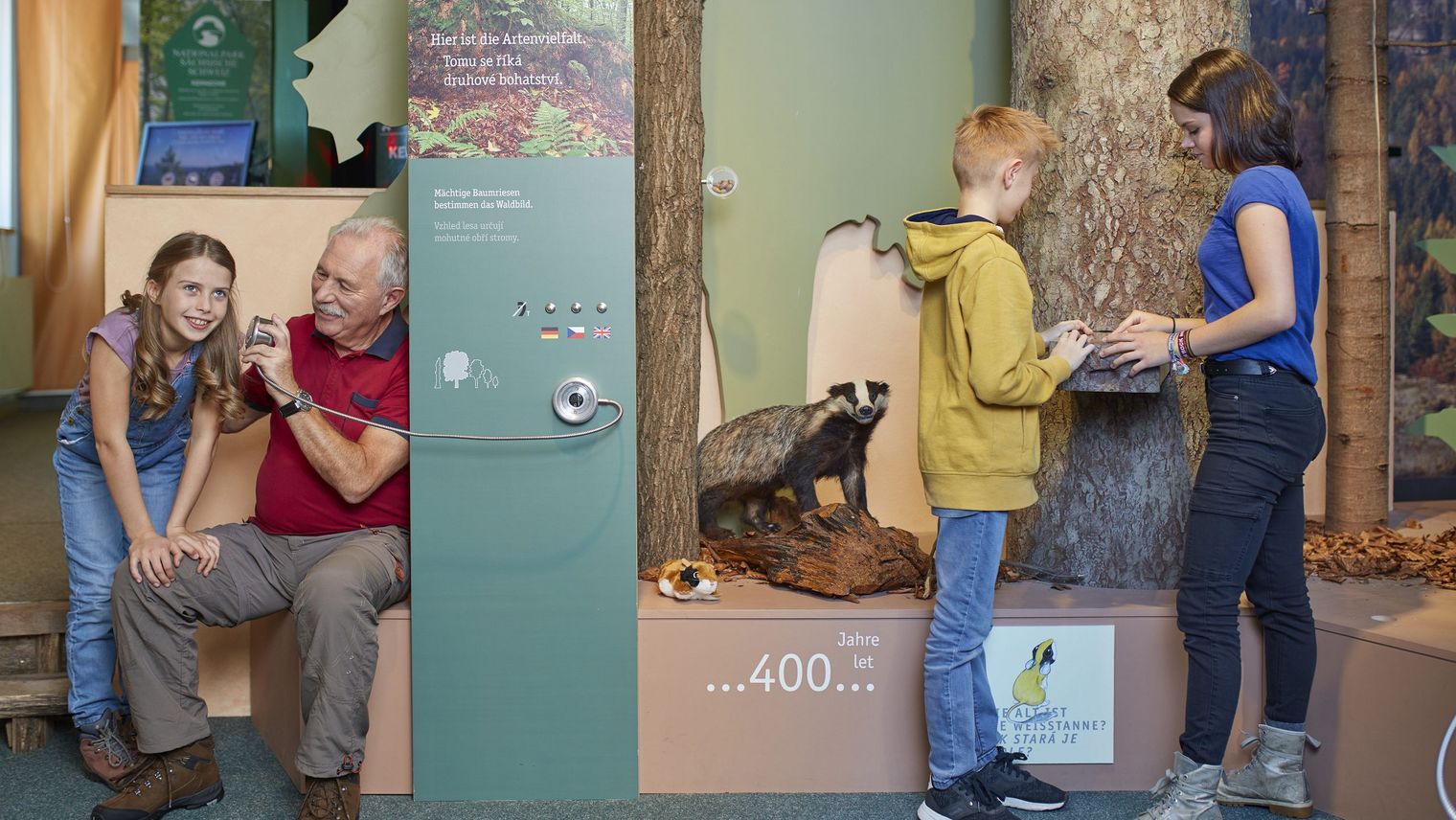 Ein älterer Herr mit einem Mädchen an einer Hörstation sowie ein Junge und ein Mädchen an einer Taststation im NationalparkZentrum Sächsische Schweiz 