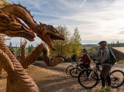 Eine Familie steht mit ihren Rädern am Rande der Blockline-Tour. Sie schauen auf Drachenfiguren aus Holz.