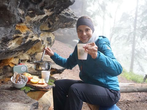 eine Frau sitzt warm angezogen unter einem Felsvorsprung und genießt ein Picknick