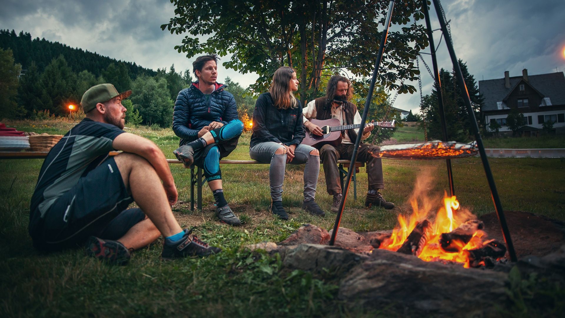 Eine Gruppe sitzt gemütlich am Lagerfeuer zusammen und ein Mann spielt ein Lied auf der Gitarre. 