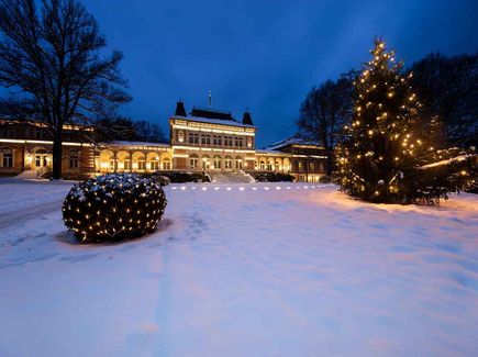 Das Bild zeigt ein weihnachtlich geschmücktes, herrschaftliches Gebäude mit einem Vorplatz. Auf diesem steht ein geschmückter und beleuchteter Weihnachtsbaum.