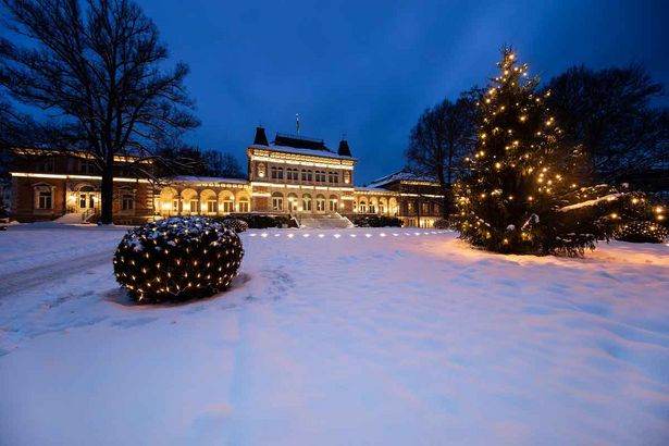 Das Bild zeigt ein weihnachtlich geschmücktes, herrschaftliches Gebäude mit einem Vorplatz. Auf diesem steht ein geschmückter und beleuchteter Weihnachtsbaum.