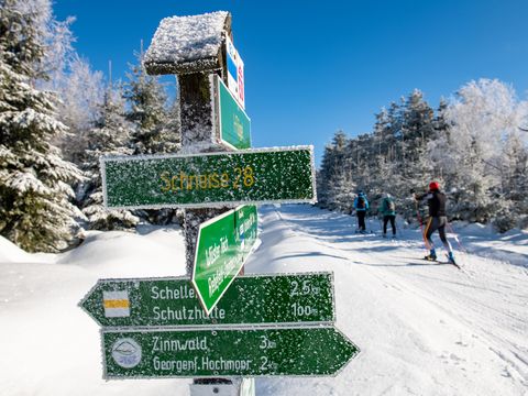 verschneite Langlaufloipe mit Sportlern und Wegweiser Schild im Vordergrund
